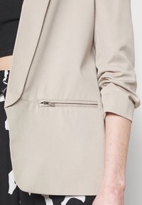 Blazer beige clair avec une texture lisse et douce, doté d'une poche zippée sur le côté. Manches retroussées et coupe ajustée.