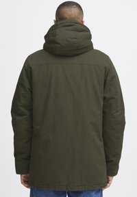 Chaqueta con capucha de color verde oliva, con un corte relajado, tela gruesa y costuras visibles. La parte trasera tiene un diseño sin costuras y sin adornos.