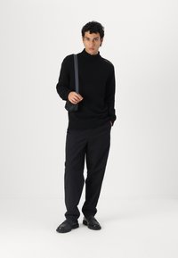 Μαύρο πλεκτό πουλόβερ τύπου turtleneck με ριγωτό τελείωμα, σε συνδυασμό με χαλαρούς μαύρους παν trousers και μαύρα παπούτσια. Το μοντέλο κρατά μία μικρή μαύρη τσάντα ώμου.