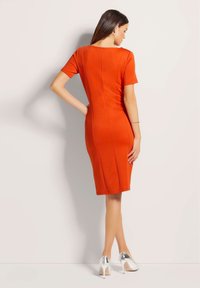 Oranje aansluitende jurk met korte mouwen, naaddetails, ronde halslijn en een rits aan de achterkant, gecombineerd met zilveren hoge hakken.