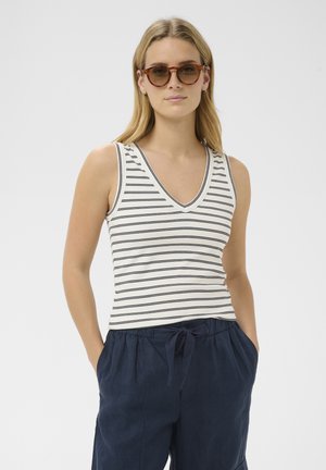 Femme portant des lunettes de soleil écaille de tortue, un haut blanc sans manches à col en V avec des rayures noires, et un pantalon bleu marine à cordon avec les mains dans les poches.