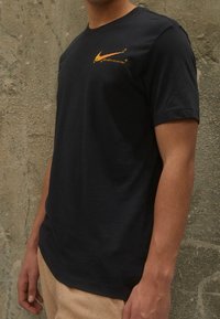 Svart t-shirt i bomull med korta ärmar, med en gul Nike-logotyp på bröstet. Slät textur, klassisk rundhalsad design.