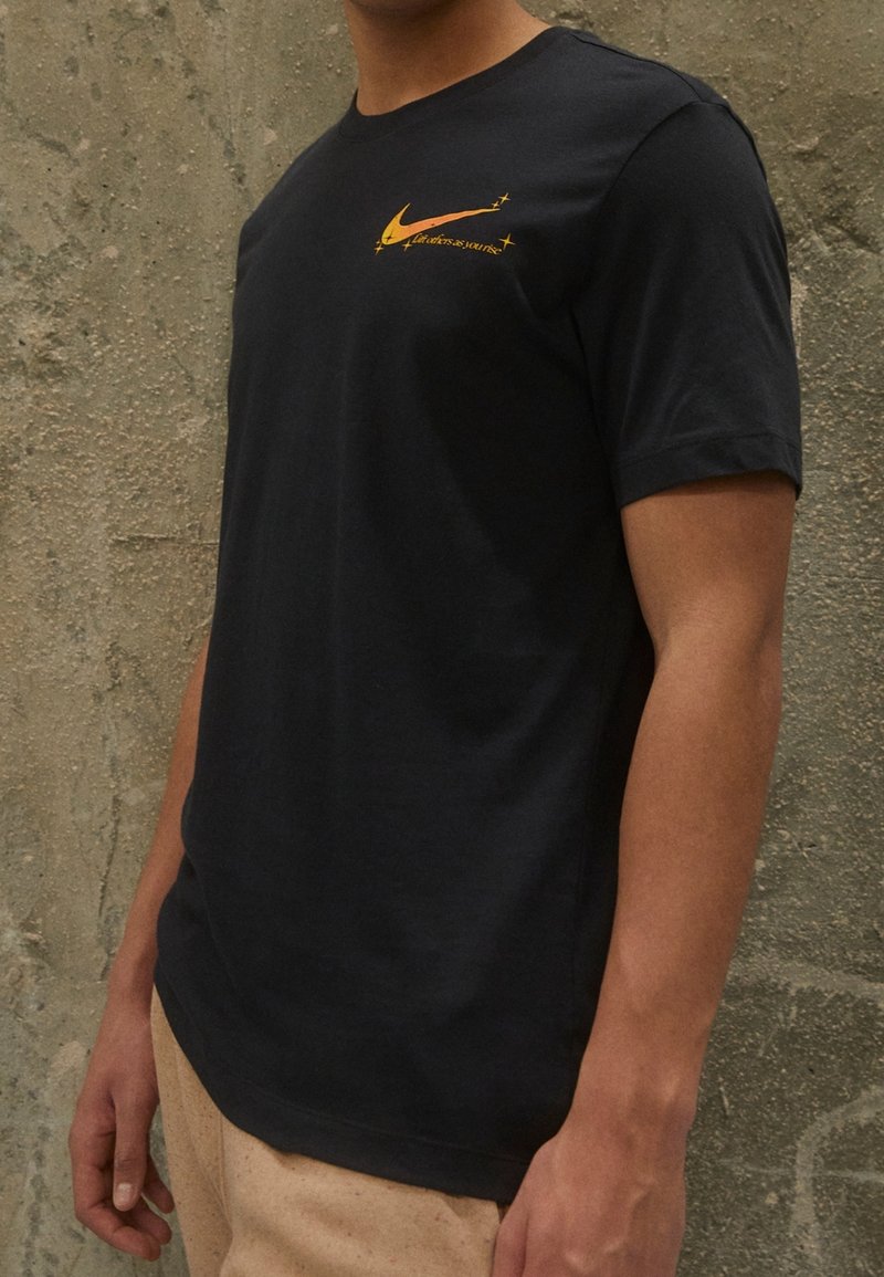 Svart t-shirt i bomull med korta ärmar, med en gul Nike-logotyp på bröstet. Slät textur, klassisk rundhalsad design.