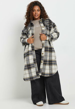 Camicia lunga a scacchi in bianco, blu e verde oliva, con chiusura a bottoni, grandi tasche e una texture morbida e pelosa. Abbinata a pantaloni scuri.