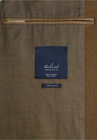 Intérieur d'un manteau en mélange de laine brun avec une étiquette marine indiquant "tailored MNG MAN", taille moyenne pour EUR-USA-MEX, fabriqué en Chine.