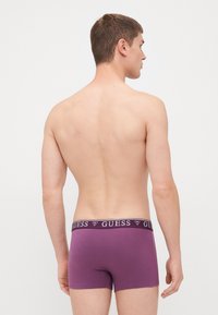 Violet vīriešu boksera apakšbikses ar melnu jostu, uz kuras ir "GUESS" logo. Mīksts audums ar pieguļošu dizainu un īsu kāju garumu.