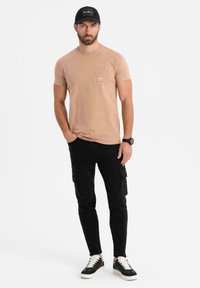 Ljusbeige t-shirt med bröstficka, bärs tillsammans med svarta cargobyxor med sidofickor, samt svarta sneakers med vita accenter.