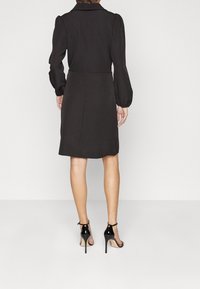 Robe noire avec une silhouette ajustée, des manches bouffantes et un col. Associée à des escarpins noirs à lanières, mettant en valeur des jambes nues.