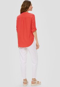 Rotes Langarmshirt mit hochgerollten Ärmeln, abgerundetem Saum und Rückenschlitzdetails. Kombiniert mit weißen, leichten, schmal zulaufenden Hosen und hellbraunen Sandalen.