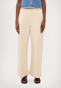 Pantalones de mezclilla beige de pierna ancha con cintura alta. Detalles notables incluyen un dobladillo sin terminar y costuras en contraste. Sandalias negras de tiras mostradas.
