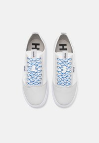 Une paire de baskets basses blanches avec des lacets à motifs bleus et blancs, des onglets de tirage de marque et un logo "H" noir sur les semelles intérieures.