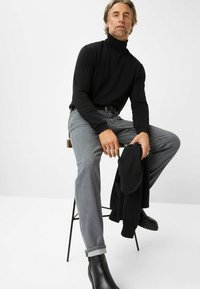 Schwarzer Rollkragenpullover, graue Jeans mit hochgekrempelten Bündchen und schwarze Stiefeletten. Person sitzt auf einem Hocker und hält einen schwarzen Pullover. Schlichter Hintergrund.