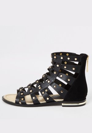 Sort sandalstøvle med korsede remme og gyldne nitter, med lynlåslukning, flad sål og en suede-detalje. Guldtrim på hælen.