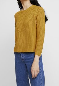 Senfgelbes Strickpullover mit strukturiertem Muster, rundem Ausschnitt und Dreiviertelärmeln, kombiniert mit blauen Jeans.