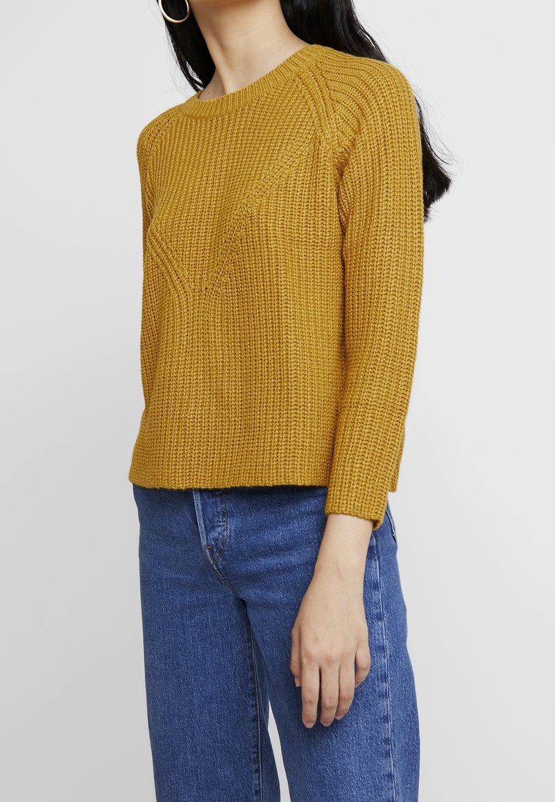 Senfgelbes Strickpullover mit strukturiertem Muster, rundem Ausschnitt und Dreiviertelärmeln, kombiniert mit blauen Jeans.