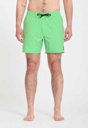 Homme portant un short de bain vert vif avec un cordon de serrage noir, debout pieds nus devant un fond blanc uni.