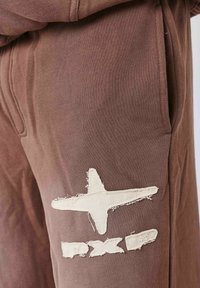 Joggers in cotone marrone con un'applicazione strappata di colore crema, raffigurante una stella e un testo sulla coscia sinistra. Tessuto morbido, vestibilità regolare.