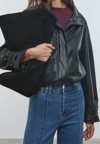 Mustat nahkatakki, jossa on hieman ylipitkä malli, yhdistettynä korkeavyötäröisiin sinisiin farkkuihin. Pitelee mustaa mokkakukkaroa.