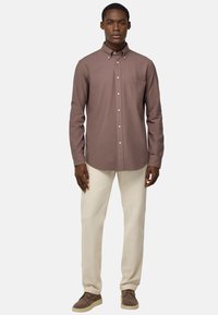 Camicia marrone con bottoni, dotata di una tasca sul petto, indossata sopra pantaloni color crema e scarpe marroni, caratterizzata da una vestibilità rilassata e una texture liscia.