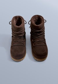 Barna szarvasbőr csizma puha béléssel, lekerekített orral, fűzős elülső résszel és gumitalppal. Nincsenek rajta hangsúlyos minták vagy díszítőelemek.