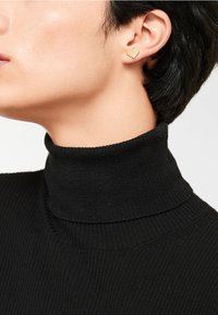 Guld hjärtformade stiftörhängen med geometrisk design, bärs av en modell i en svart polotröja. Texturen är slät och polerad.