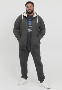 Felpa grigia con zip e cordini crema, abbinata a pantaloni della tuta coordinati. Sneakers bianche completano l'outfit, con una t-shirt scura stampata indossata sotto.