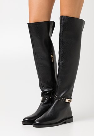Botas de cuero negras sobre la rodilla con acabado texturizado. Cuenta con un cierre lateral y una correa con tachuelas en el tobillo con herrajes dorados. Suela plana.