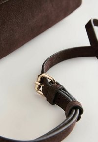 Sangle en daim marron foncé avec une boucle dorée attachée à un sac assorti sur une surface blanche.