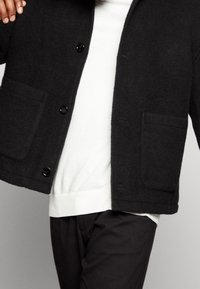 Giacca di lana nera con colletto rotondo, tasche frontali e bottoni neri. Indossata sopra un maglione bianco, abbinata a pantaloni scuri.