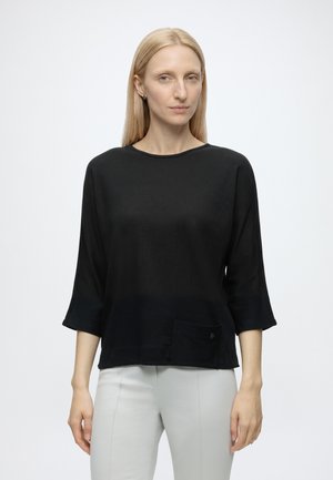 Femme aux cheveux blonds raides portant un haut noir ample à manches trois-quarts et un pantalon gris clair, debout devant un fond uni.