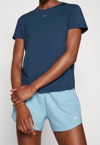 Camiseta atlética de manga corta azul marino de Nike con cuello redondo, combinada con pantalones cortos azul claro que presentan un logo blanco en el lateral. Textura de tela suave.
