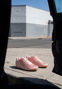 Paire de baskets roses posées sur un trottoir ensoleillé près d'une porte de voiture ouverte avec un bâtiment gris et blanc en arrière-plan.