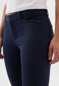 Personne portant un pantalon bleu marine foncé ajusté et une chemise blanche rentrée, debout avec les mains détendues le long du corps sur un fond clair uni.