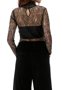 Schwarzes Spitzentop mit einem hohen Kragen, langen Ärmeln und einem Schlüsselloch-Rücken, kombiniert mit einem einfarbigen schwarzen Stoff an der Taille.