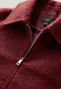 Veste bordeaux à texture douce avec fermeture éclair argentée partiellement ouverte, dévoilant une doublure intérieure lisse et une étiquette de vêtement Next noire.