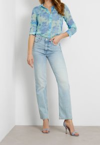 Kobieta ubrana w prześwitującą niebieską koszulę w kwiaty zapinaną na guziki, jasnoniebieskie jeansy z wysokim stanem i prostą nogawką oraz jasnoniebieskie buty na obcasie z odkrytymi palcami.