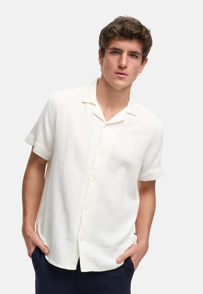 Camisa de manga corta abotonada en tejido blanco texturizado, con cuello con muesca y sutil detalle de logo, llevada con las manos en los bolsillos.