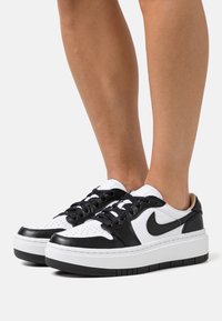 Baskets noir et blanc avec une tige en cuir, bout perforé, lacets noirs plats et une épaisse semelle blanche avec un dessous noir texturé.