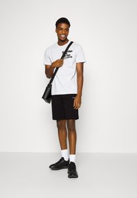 Hvit bomull t-skjorte med grafisk trykk, kombinert med svarte shorts. Svart crossover-veske og svarte sneakers, supplert med hvite ankelsokker.