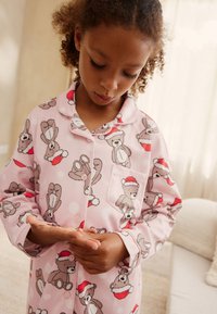 Pink pajama shirt featuring teddy bear patterns wearing Santa hats. Made of soft fabric with a button-up design and patch pocket.