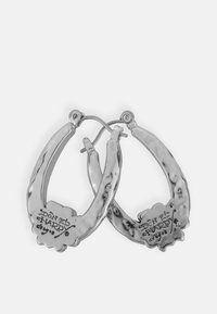Orecchini a cerchio in argento con superficie testurizzata, caratterizzati da dettagli del marchio incisi sul retro. Il design è aperto e di forma ovale.