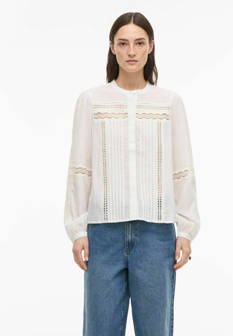 Blouse blanche avec des détails découpés complexes et des rayures verticales, dotée de manches longues, d'un col rond et d'une fermeture à boutons.