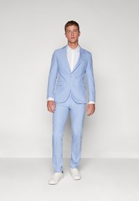 Svetlomodrý oblek so slim fit strihom, ktorý má jednoradový sak a nohavice s plochým predným dielom, skombinovaný s bielou košeľou a bielymi teniskami.