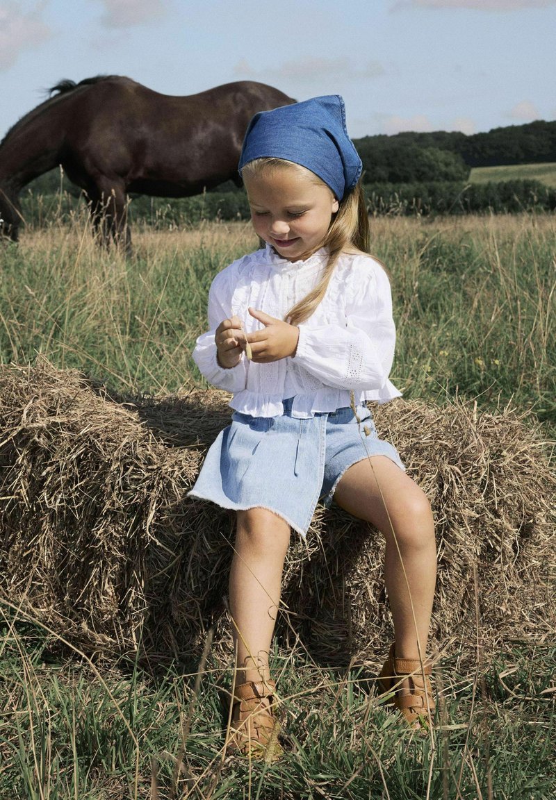 Ragazza in una camicia bianca con volant e gonna di jeans blu seduta su un mucchio di fieno, mentre tiene della paglia. Indossa un foulard blu e stivali marroni.
