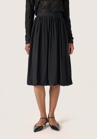 Gonna nera di lunghezza al ginocchio con vita arricciata e tessuto fluido, abbinata a scarpe nere a punta. Texture liscia, design semplice.