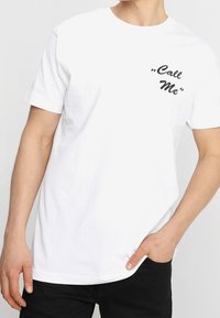 Witte katoenen t-shirt met korte mouwen, een ronde hals en een zwarte tekstprint die "Bel Me" zegt op de bovenste linkerzijde.