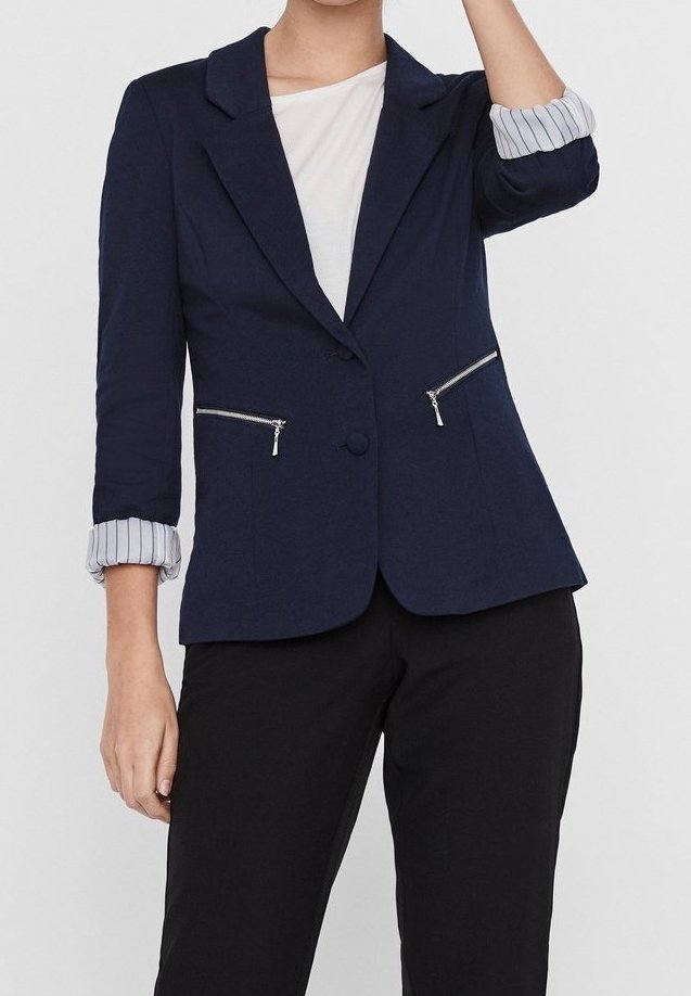 Personne portant un blazer bleu marine avec les manches rayées retroussées, une chemise blanche en dessous et un pantalon noir, debout avec un bras levé.