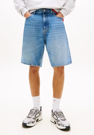 Person trägt hellblaue Jeansshorts, weiße Sportsocken und weiß-graue Turnschuhe und steht mit den Händen in den Taschen.