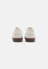 Weiße Wildledersneaker mit einer strukturierten braunen Gummisohle, die sichtbare weiße Markenbeschriftung an der Ferse und ein schlichtes Design aufweisen.