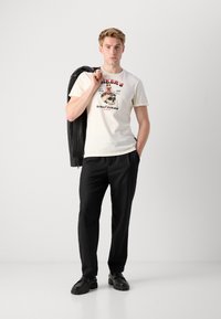 Camiseta crema con estampado gráfico, chaqueta ligera negra sobre el hombro, pantalones negros y zapatos negros sobre un fondo neutro.